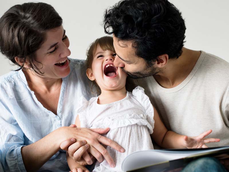 Mom and dad laughing with a child on their lap reading a book
